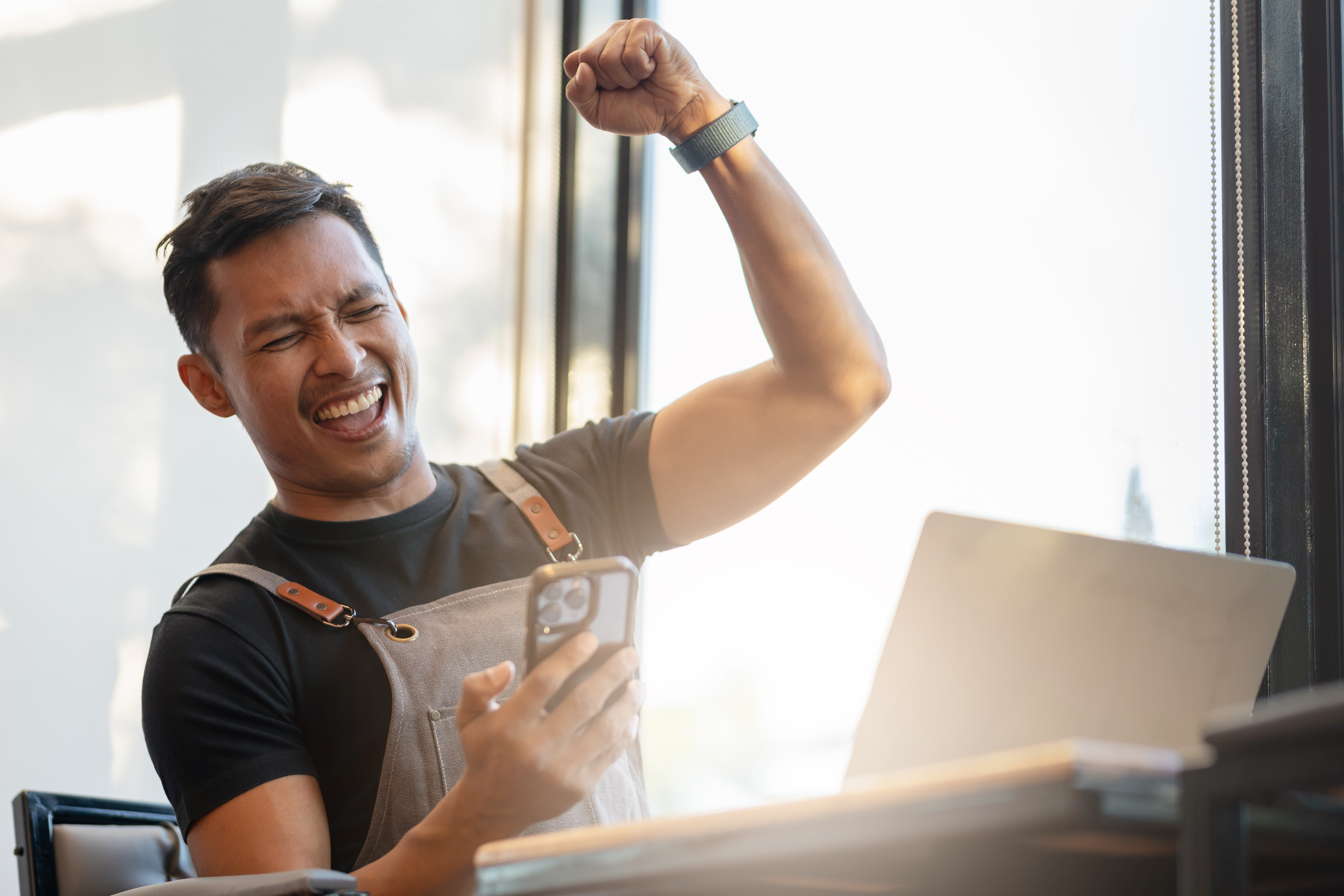 A man in an apron cheers with a raised fist while looking at his smartphone, with a laptop nearby.