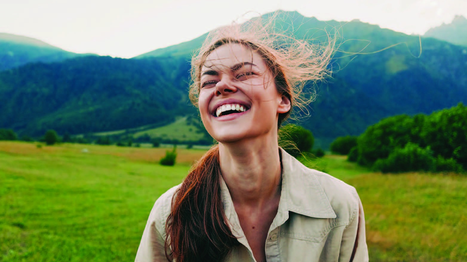 A young woman laughing joyfully outdoors, hair blowing in the wind, with green mountains and meadows in the background.