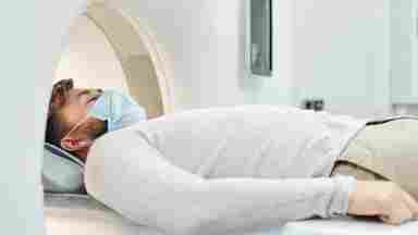 Young male in casual wear and protective mask about to receive a scan in a large MRI machine.