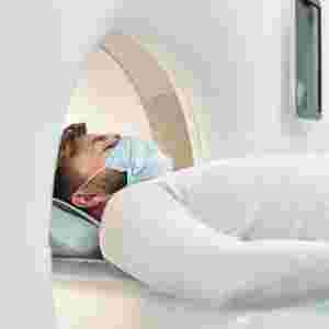 Young male in casualwear and protective mask lying on long couchette and moving into large MRI imaging equipment for examination. Scan. 