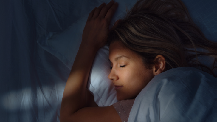 A young woman with long brown hair sleeping peacefully on a white pillow in a dimly lit bedroom at night.