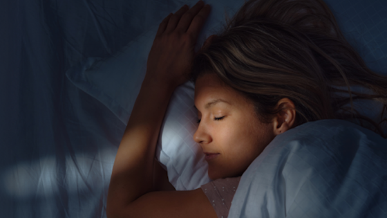 A young woman with long brown hair sleeping peacefully on a white pillow in a dimly lit bedroom at night.