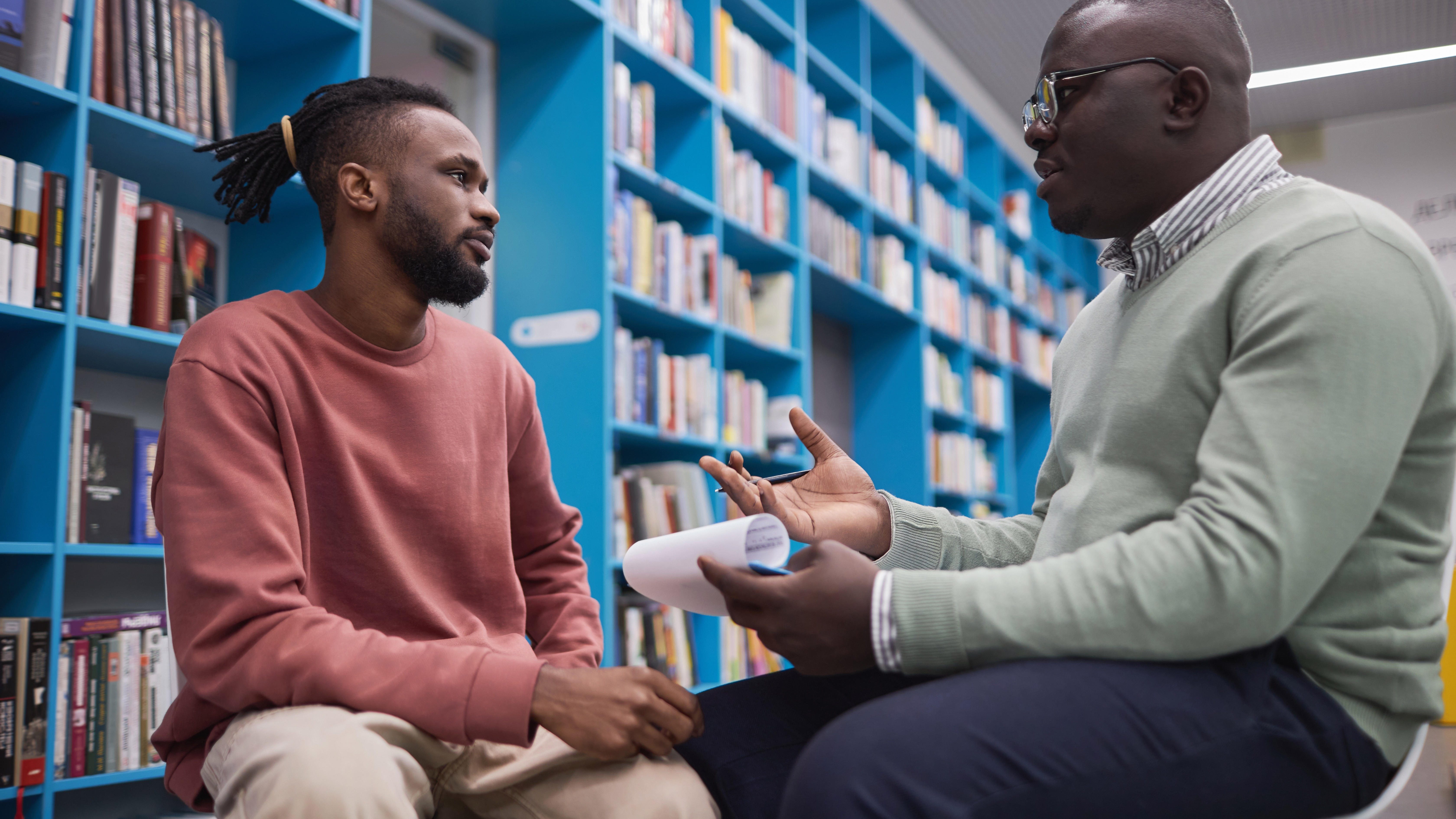 Two Black men having a serious conversation in a library, the younger man in a pink sweater listening, the other, perhaps a teacher, holding papers with notes.