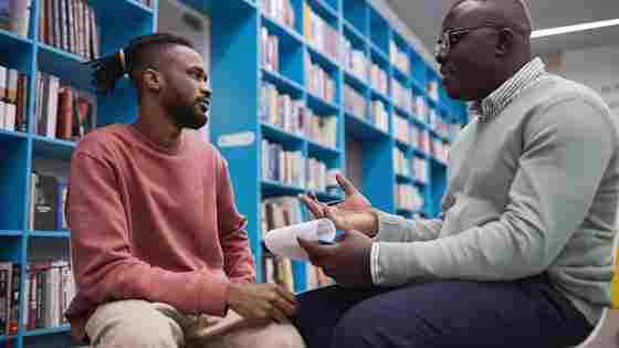 Two Black men having a serious conversation in a library, the younger man in a pink sweater listening, the other, perhaps a teacher, holding papers with notes.