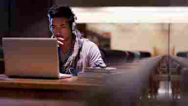 A college student wars headphones while working on their laptop in a quiet library.