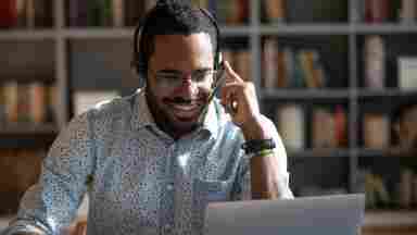 A happy person of color is wearing a headset on his computer.