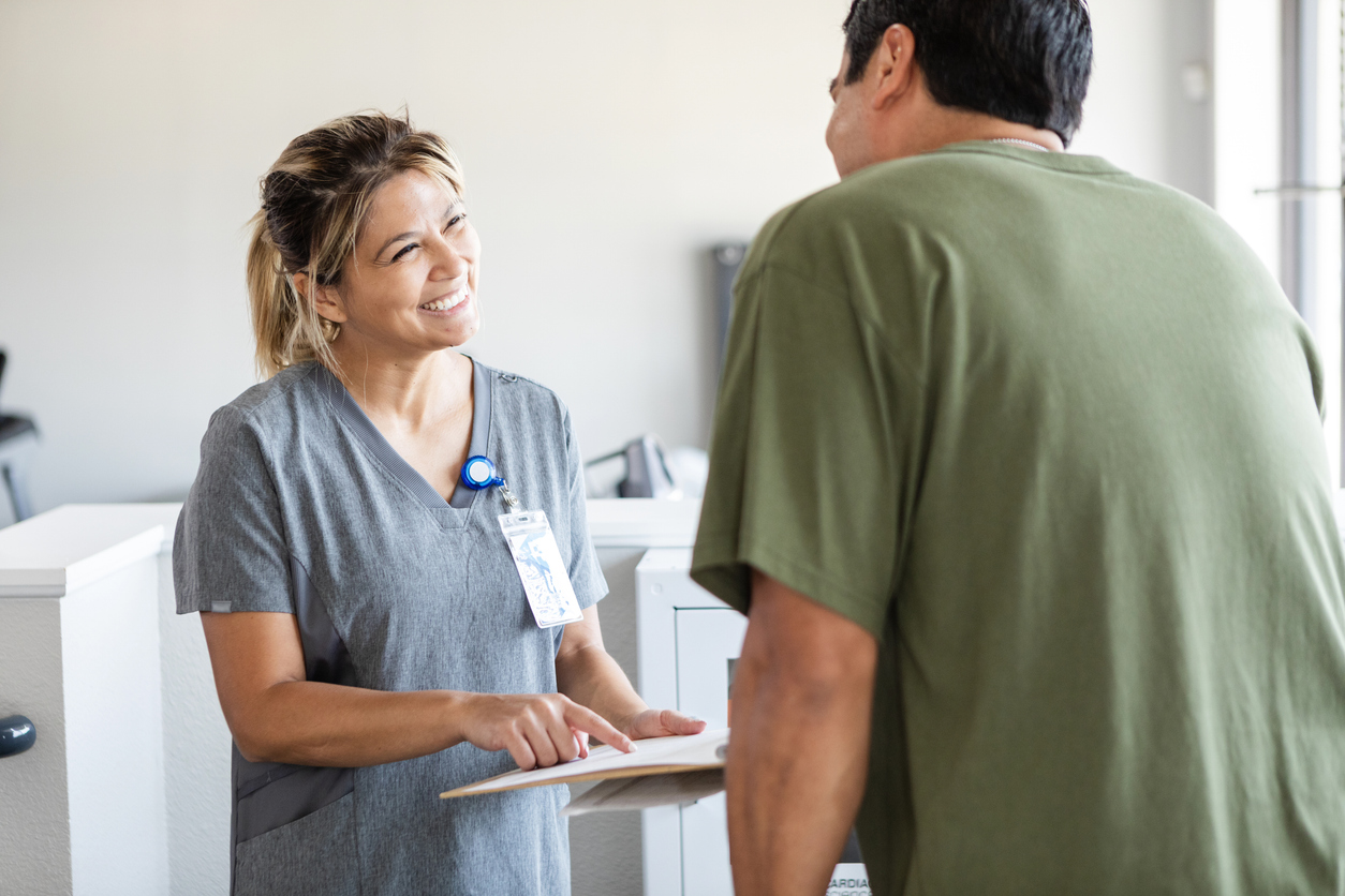 Smiling healthcare worker in gray scrubs with ID badge speaking to a patient in green shirt in a medical office.