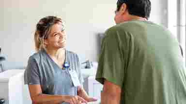 Smiling healthcare worker in gray scrubs with ID badge speaking to a patient in green shirt in a medical office.