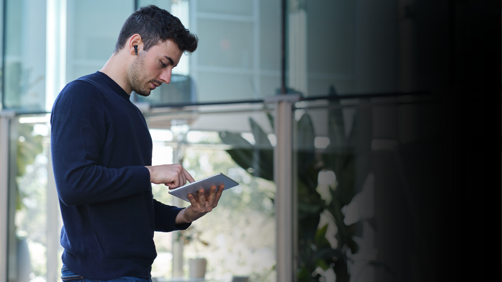A young man in a navy sweater uses a tablet outdoors near a modern glass building, wearing a wireless earbud.