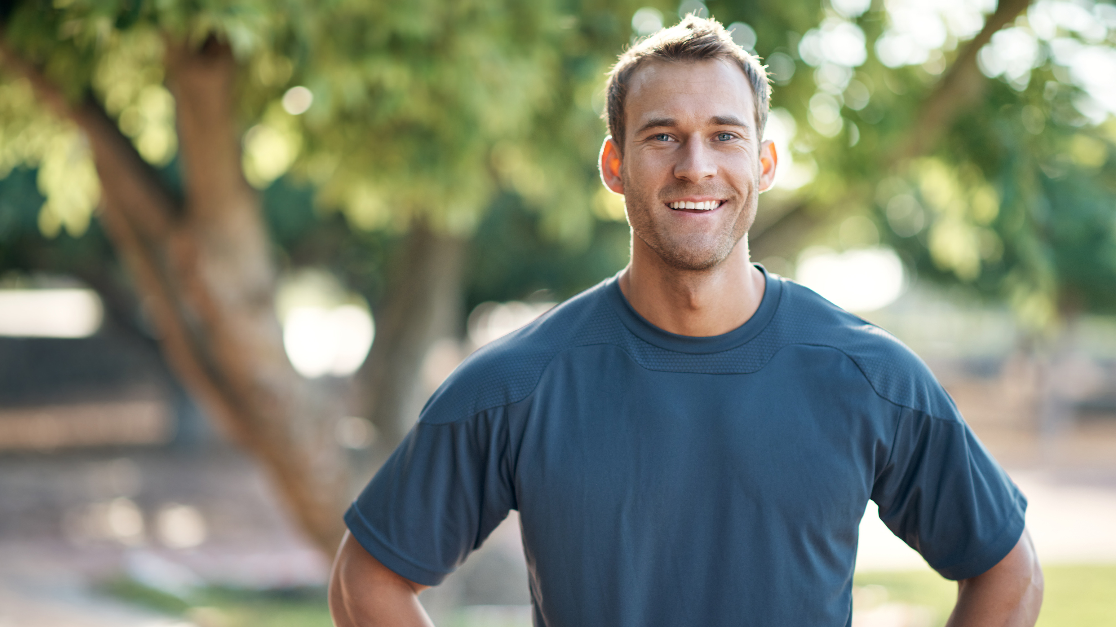 A man wearing a blue shirt stands outdoors on a sunny day; his hands are on his hips and he is smiling at the viewer.