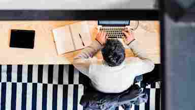 A person sits at a wooden desk and types on a small laptop.