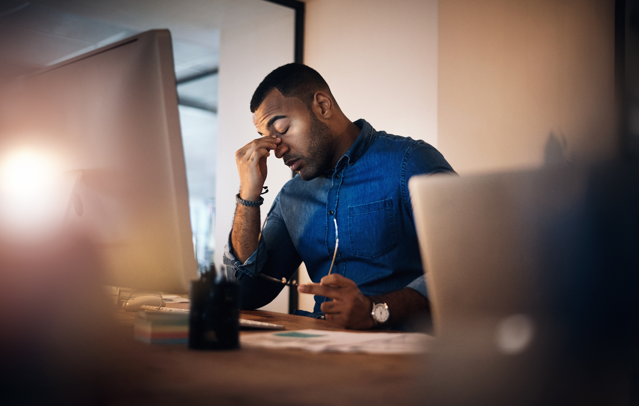 A young, Black businessman in a blue shirt looking stressed out while working in the office late at night.