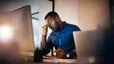 A young, Black businessman in a blue shirt looking stressed out while working in the office late at night.