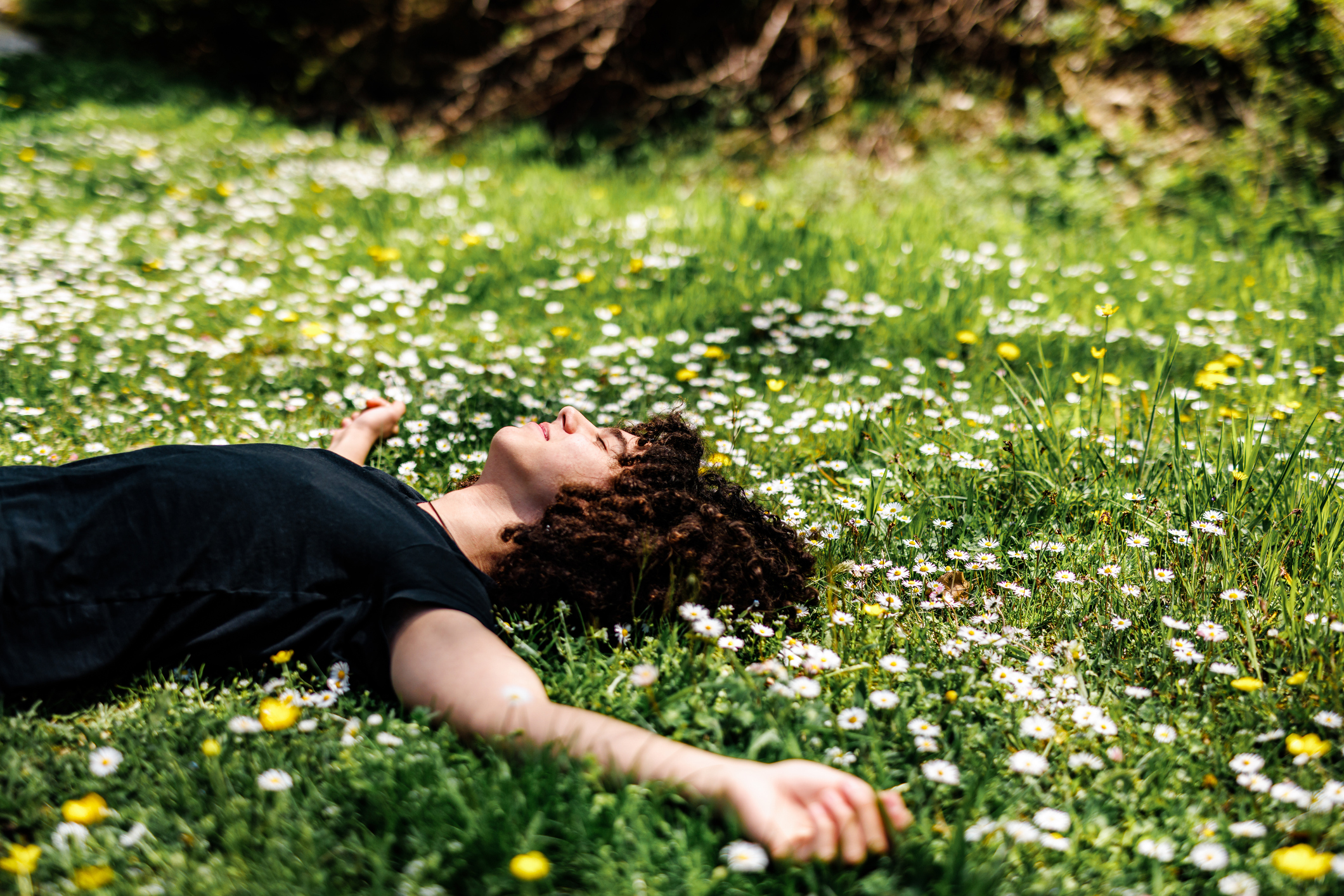 A person with curly hair in a black shirt lies peacefully on a green meadow filled with white and yellow wildflowers.