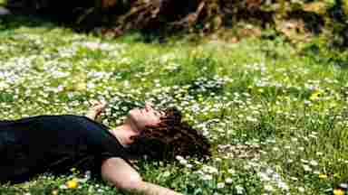 A person with curly hair in a black shirt lies peacefully on a green meadow filled with white and yellow wildflowers.