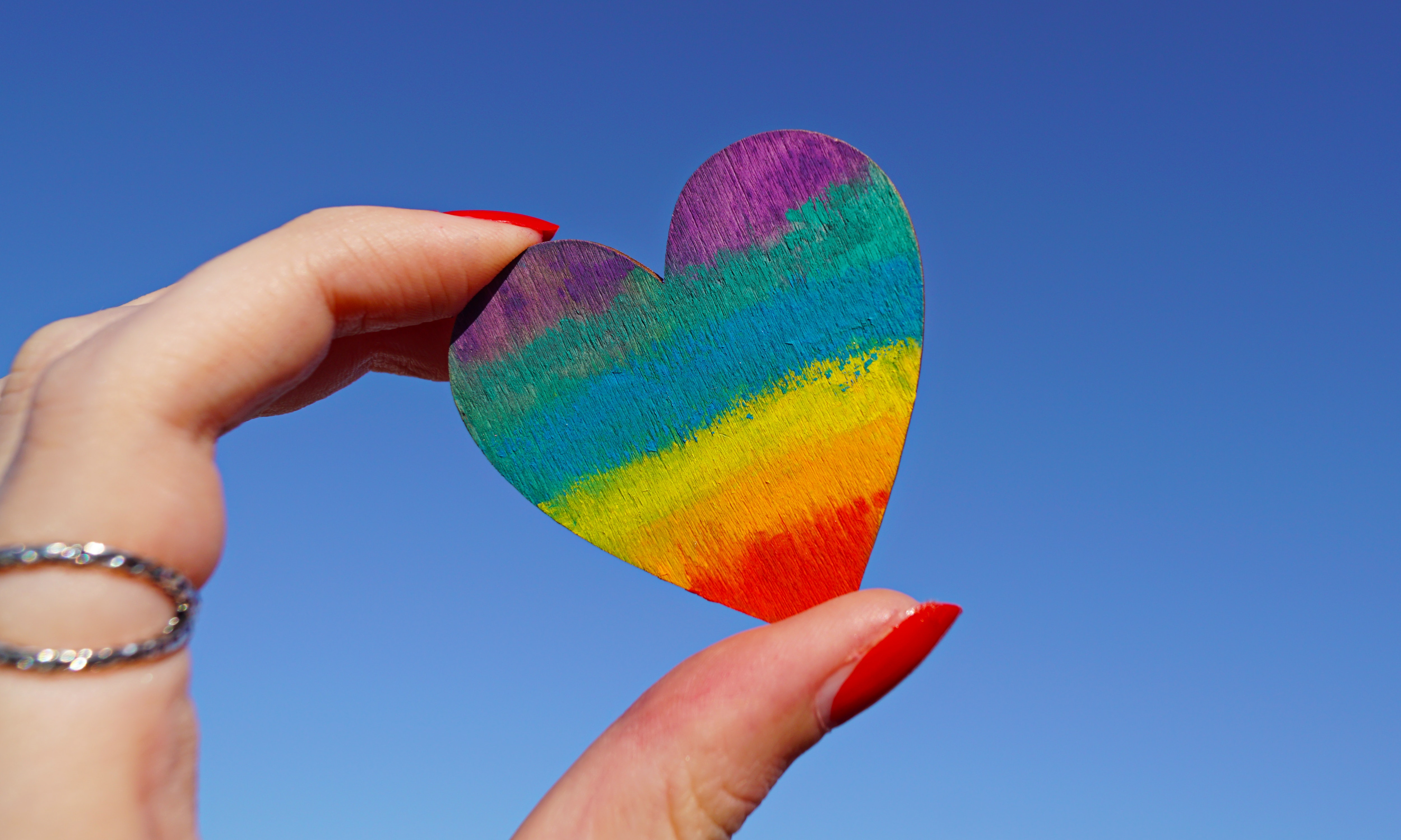 A hand holds a rainbow heart against a bright blue sky.