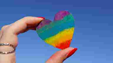 A hand holds a rainbow heart against a bright blue sky.