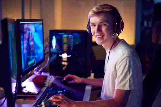 A happy young male gamer with headphones sits in front of the computer and smiling into the camera. 