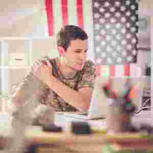 A young male soldier sits in front of a laptop with an American flag behind him. Military. First responder.