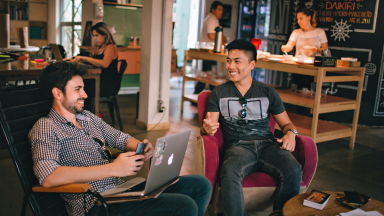 Two young professional men are having seated and smiling, having a conversation while other adults work in the background. 