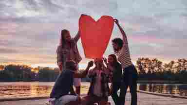 A diverse group of individuals hold a heart-shaped paper lantern on a pier at sunset.