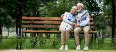 An elderly couple is sitting on a bench while holding each other in the park. Older. IStock#: 1151872303.