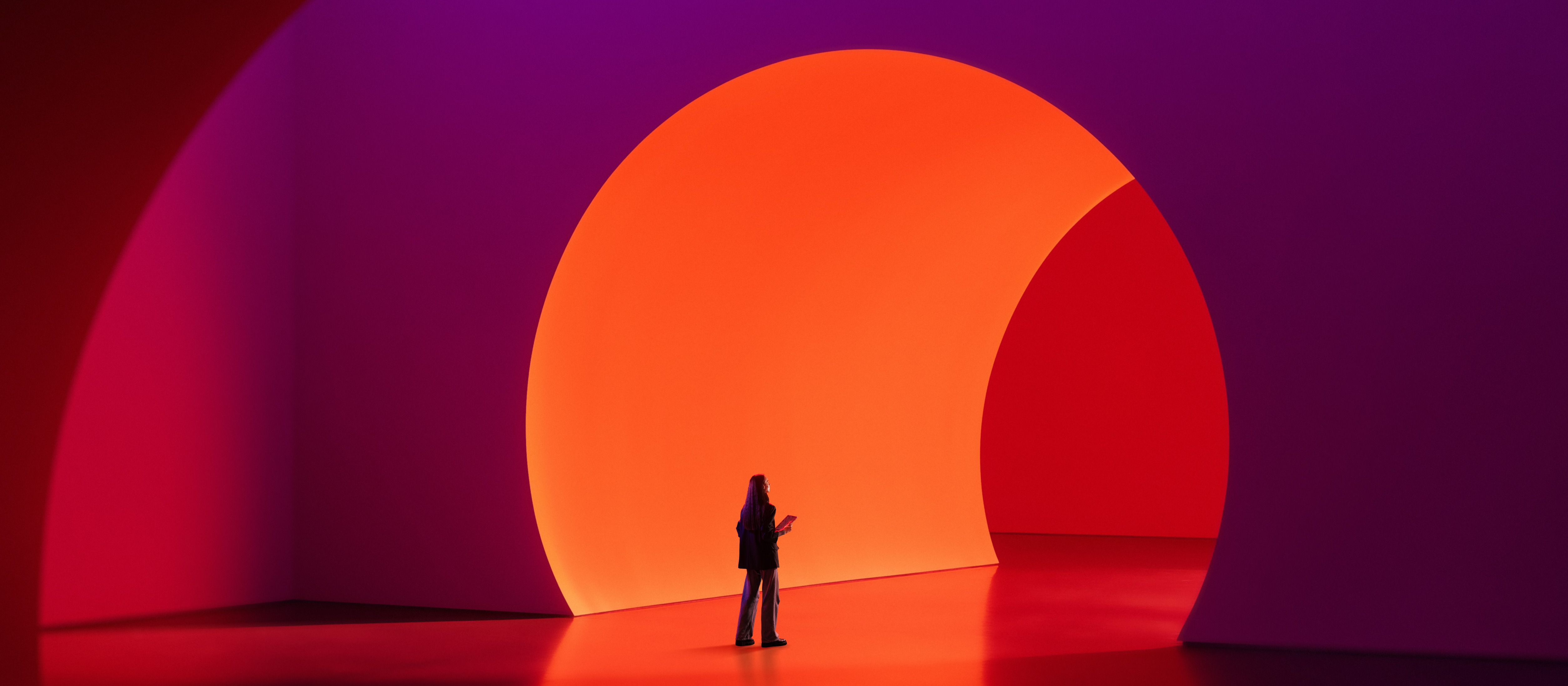 A lone person stands before a large glowing orange circular arch in a vibrant red and purple immersive art installation.