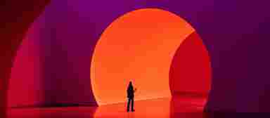 A lone person stands before a large glowing orange circular arch in a vibrant red and purple immersive art installation.