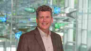 Stephen White in a brown sports coat with blue and green lights, landscape. Close-up. Chief Operating Officer