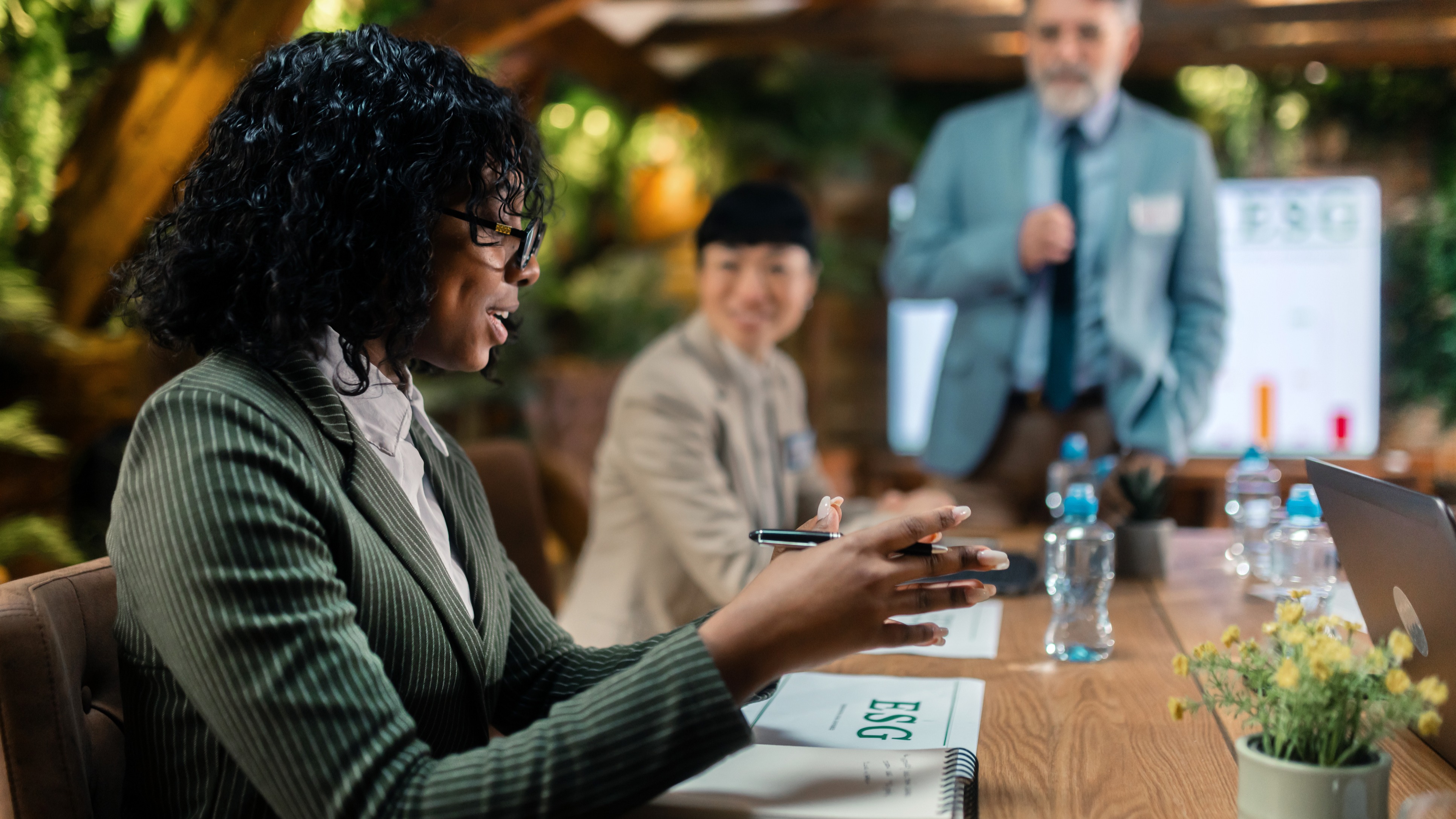A woman in a green blazer attends an ESG business meeting while a man presents at a whiteboard in a plant-filled room.