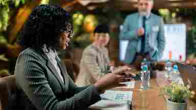 A woman in a green blazer attends an ESG business meeting while a man presents at a whiteboard in a plant-filled room.