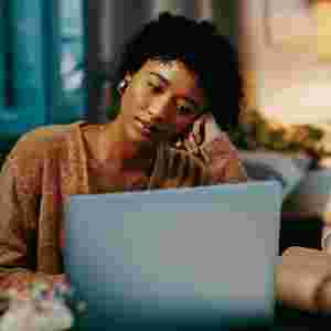 A young Black woman looking bored while working on her laptop at home in the evening.