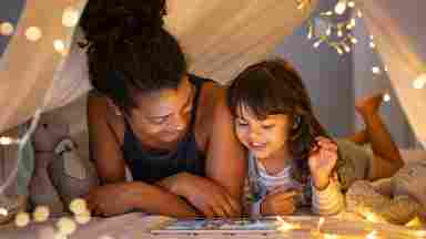 A mother and child reading in a cozy tent with hanging lights.