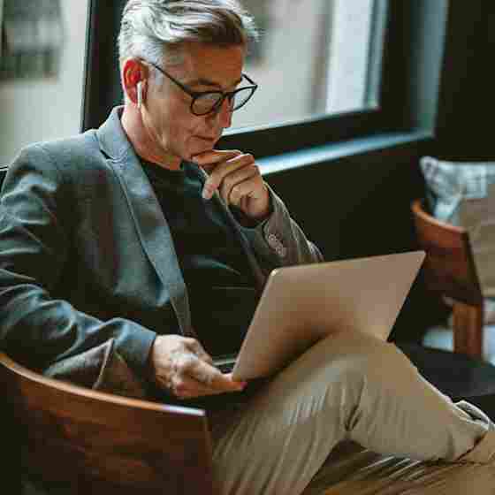 Middle-aged man with gray hair, glasses, and earbuds sits in a wooden chair, thoughtfully working on a laptop.