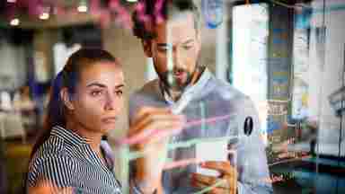 Two business people collaborating and analyzing data on a transparent whiteboard in the office.