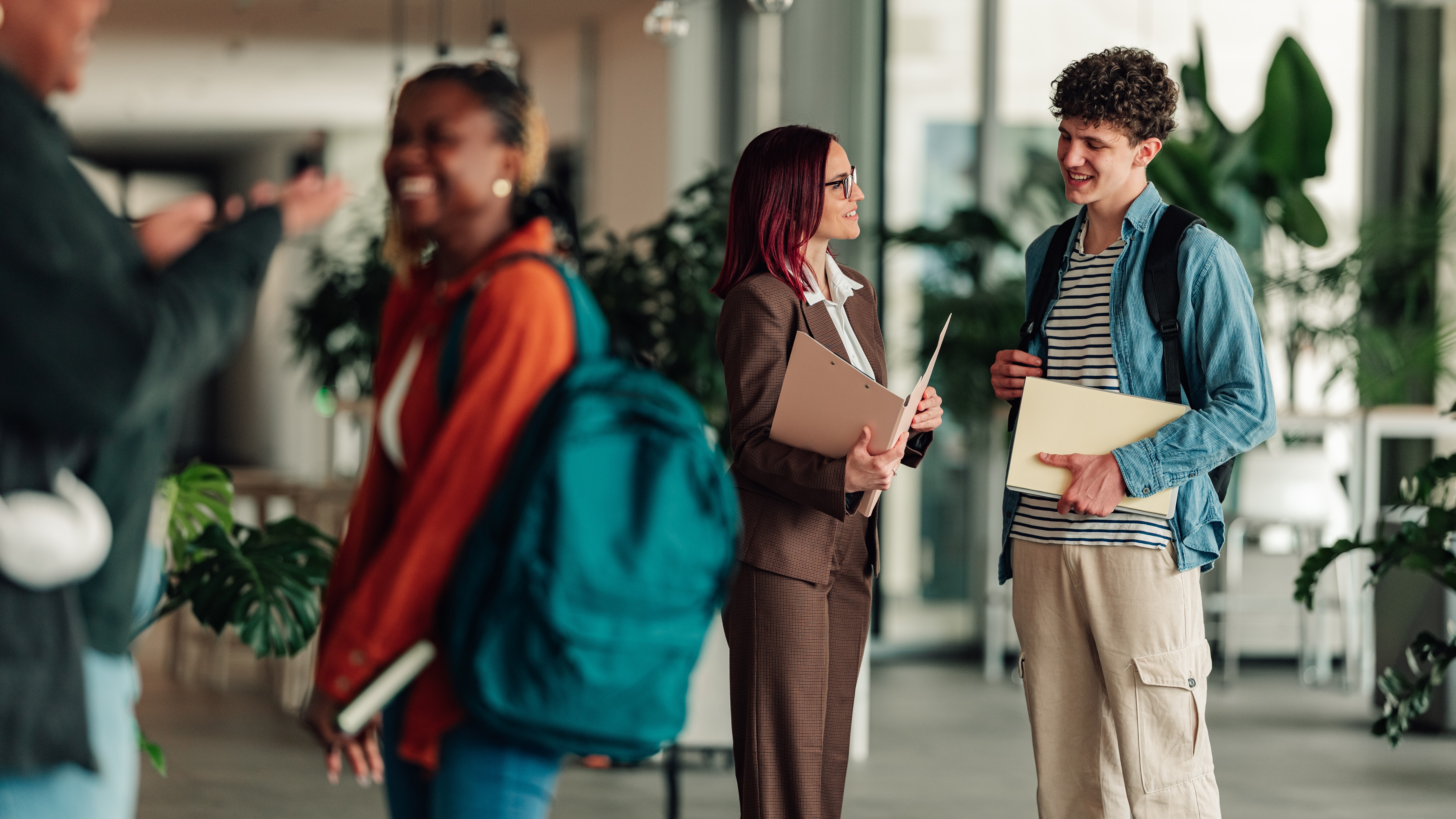 A woman in a brown suit with red hair chats with a young male student holding folders in a bright modern building.