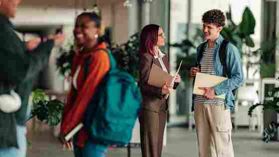 A woman in a brown suit with red hair chats with a young male student holding folders in a bright modern building.