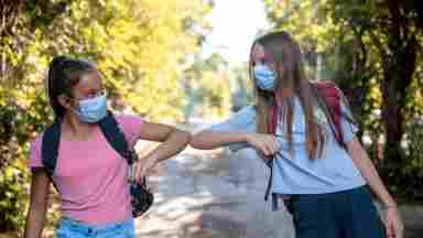 Two young female students with masks are greeting each other in a safe manner. Middle School. Youth. 