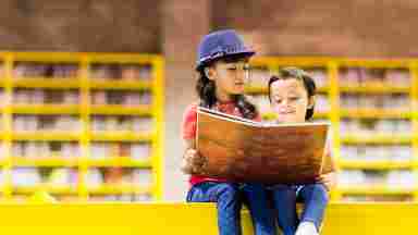 Two young children sit on a yellow ledge, reading a large book together in a yellow library setting.