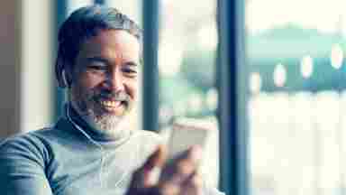 A portrait of a mature, smiling man with a beard using a smartphone and listening to music in a cafe.