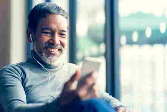 A portrait of a mature, smiling man with a beard using a smartphone and listening to music in a cafe.