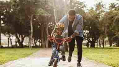 A happy father helping his smiling, helmeted child ride a bike.