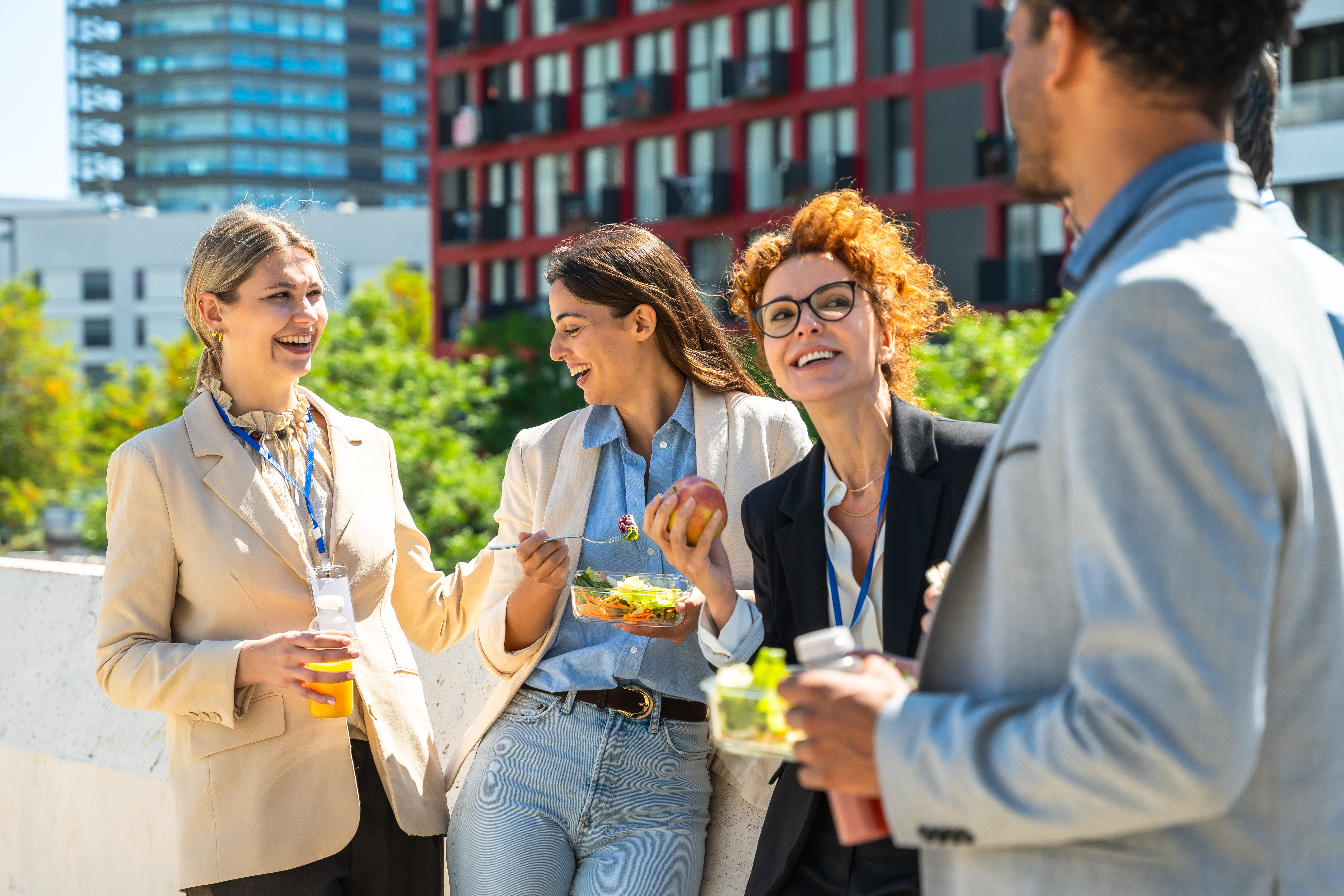 Four smiling colleagues with lanyards enjoy lunch outdoors on a sunny urban rooftop, eating salads and drinks.