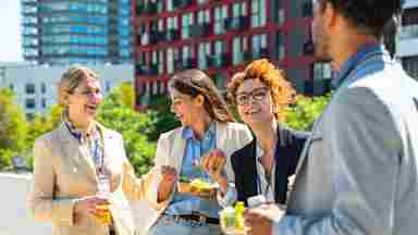 Four smiling colleagues with lanyards enjoy lunch outdoors on a sunny urban rooftop, eating salads and drinks.