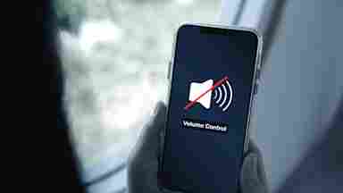 Hands holding a smartphone displaying a muted speaker icon with a red slash and the text "Volume Control" on screen.