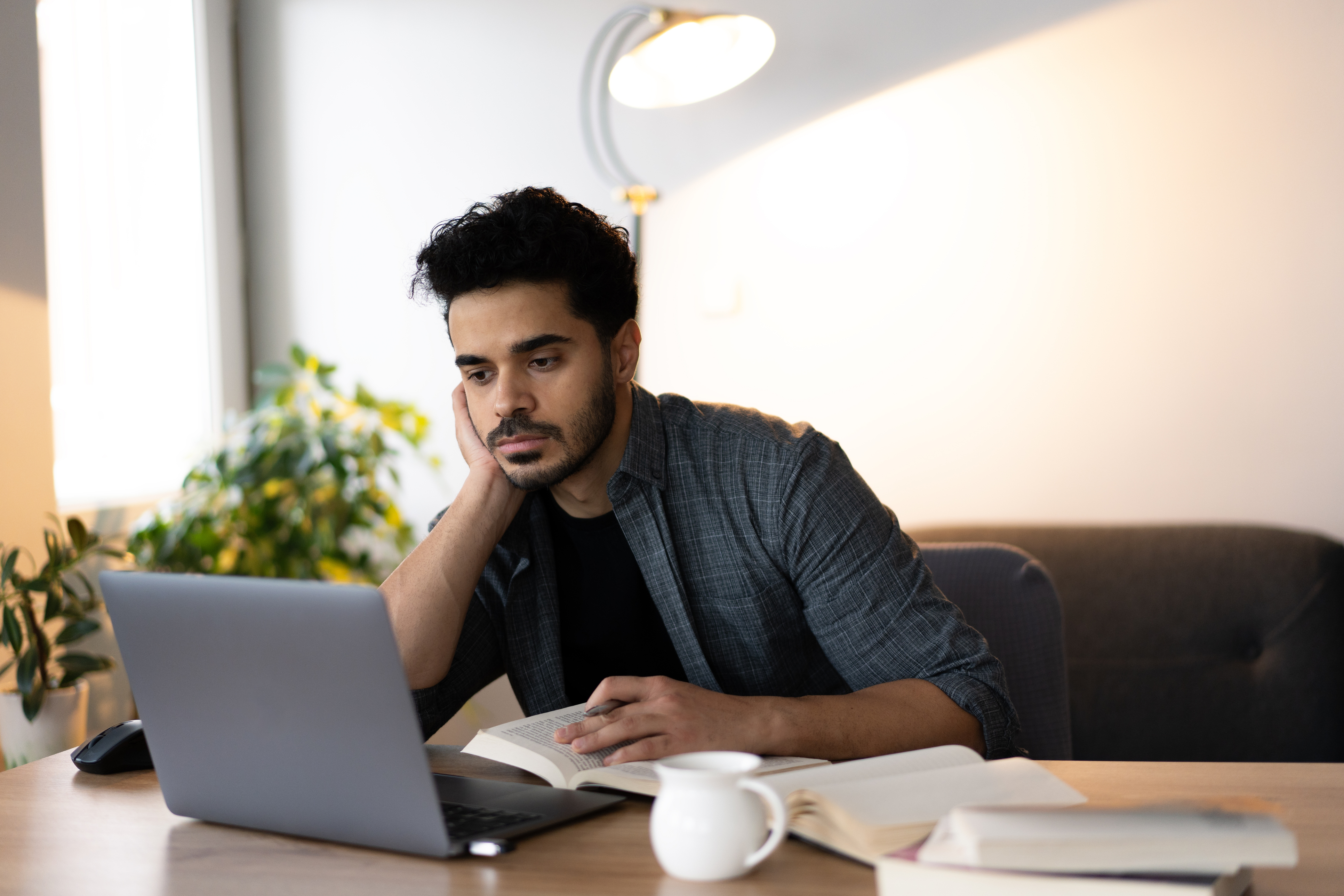 A college student focuses rests his head on his hand while reading from a laptop, with an open book and coffee mug on the desk.