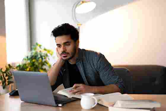A college student focuses rests his head on his hand while reading from a laptop, with an open book and coffee mug on the desk.