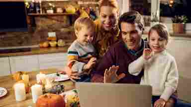 Happy parents and young children are shown using a laptop and waving while making a video call before Thanksgiving dinner in the dining room.