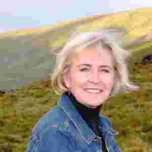A headshot of Advisory Board member Maria Conroy in front of some grassy hills and mountains; she is wearing a jean jacket and a black turtleneck.