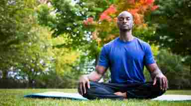 Man in a blue t-shirt sitting cross-legged on a yoga mat at a park, eyes closed